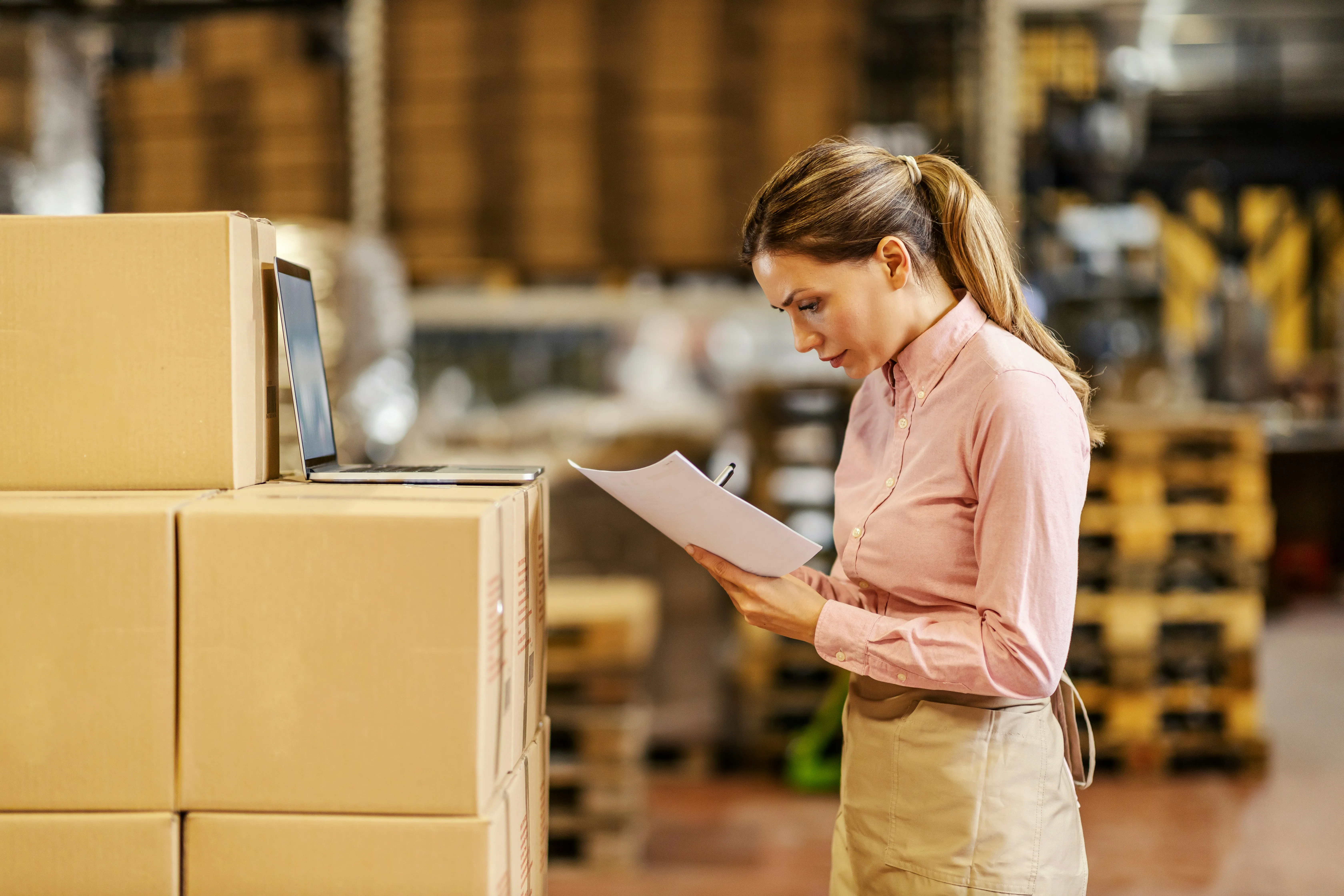 Warehouse manager reviewing paperwork and laptop among inventory boxes